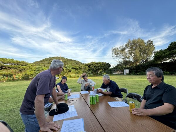 平久保半島の緑豊かな山々と海岸線を背景に、様々な年代の人々が木製のテーブルを囲んで真剣に会議している様子
